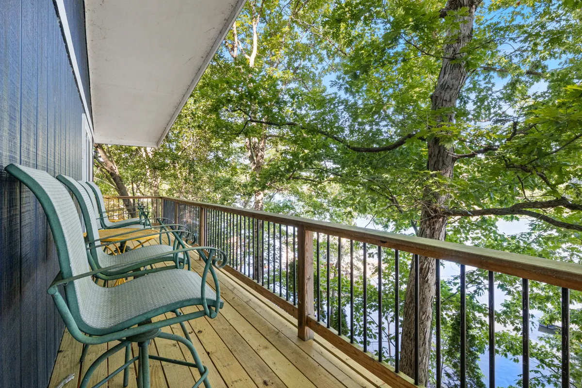 A wooden balcony with metal bar stools overlooks lush greenery and provides a clear, calming view of the lake. The inviting setup is perfect for morning coffee, birdwatching, or simply unwinding while admiring the natural surroundings.