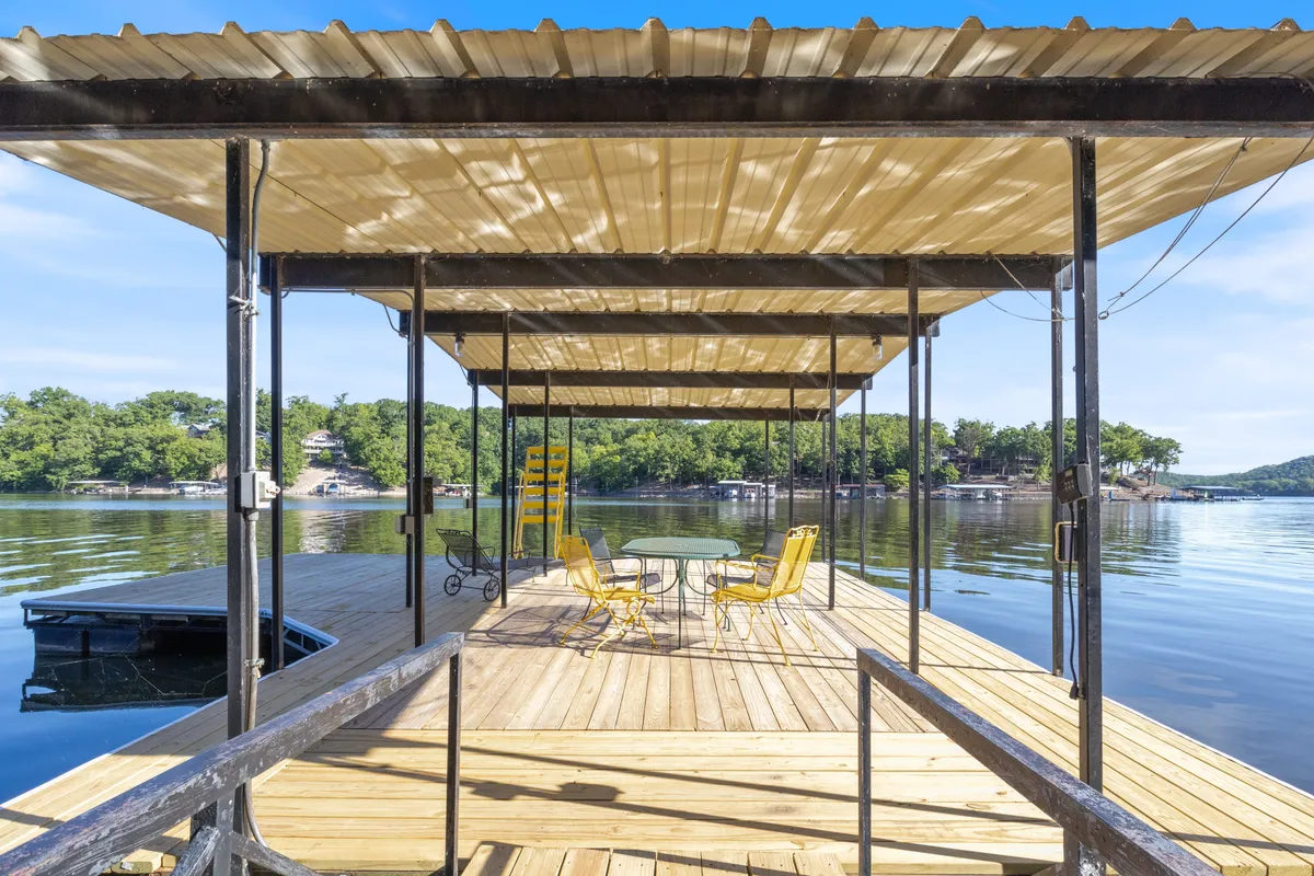 A wooden dock extends over the lake, featuring yellow chairs and a circular table. The scene evokes leisurely mornings, fishing trips, and easy access to the water for kayaking or paddle boarding. The tranquil water and inviting seating reinforce the idea of a lake adventure hub.