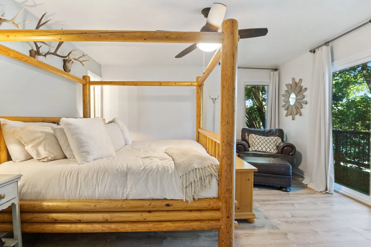 A tranquil bedroom with a four-poster bed, leather armchair, and large windows looking out onto nature. Decorative antlers and a mirror add character, enhancing the room's calm, welcoming atmosphere.