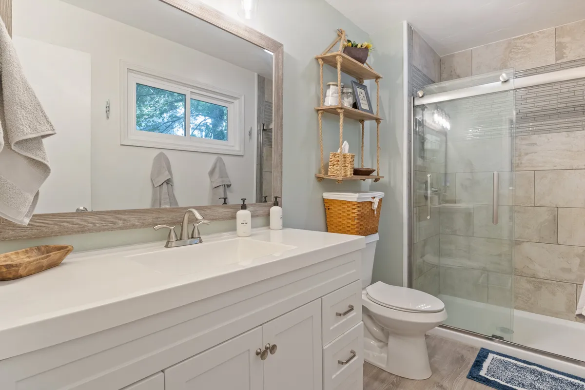 A modern bathroom featuring a spacious vanity with a sink, a large mirror, a glass shower enclosure, and neatly styled wooden shelves with tasteful decor. The space feels fresh and calming, ideal for relaxation and self-care.
