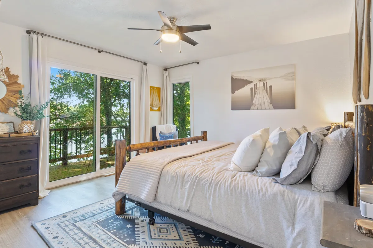 A serene and cozy bedroom featuring a plush bed adorned with decorative pillows. Large windows flood the space with natural light, offering peaceful views of the outdoors. Artwork adds a personal touch, and a wooden dresser provides practical storage.