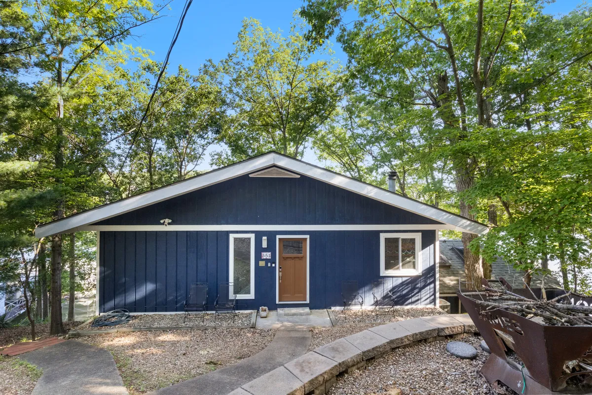 The blue exterior of the house nestled among wooded surroundings, with the front door and a hint of the outdoor living space visible. The environment feels peaceful and welcoming, suggesting easy access to both nature and local attractions.