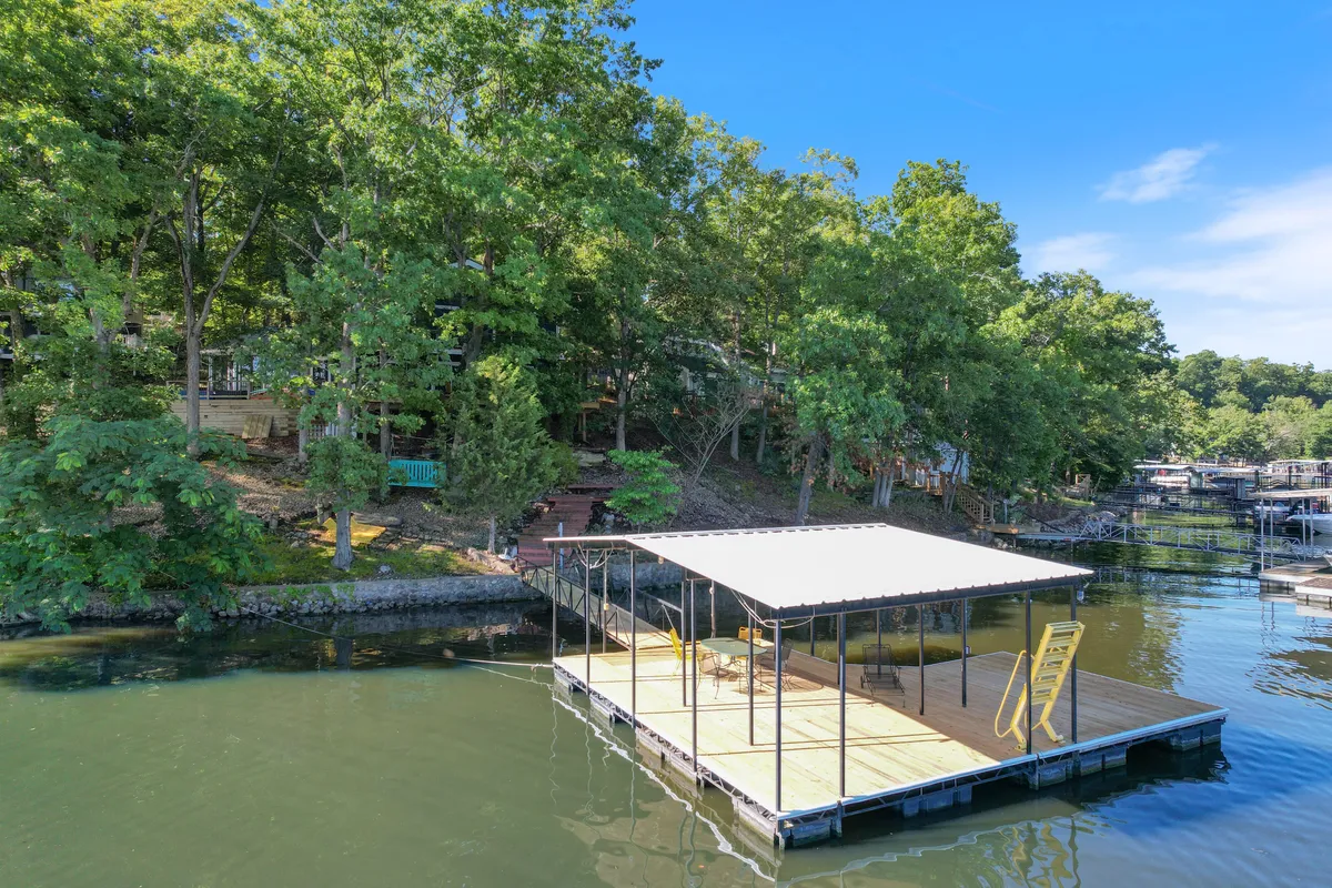 A serene lakefront scene shows a private dock extending over tranquil water, surrounded by lush trees. Comfortable seating on the dock invites fishing, relaxing, or enjoying lake views. Steps leading up to the property are visible, reinforcing the connection between home and water.