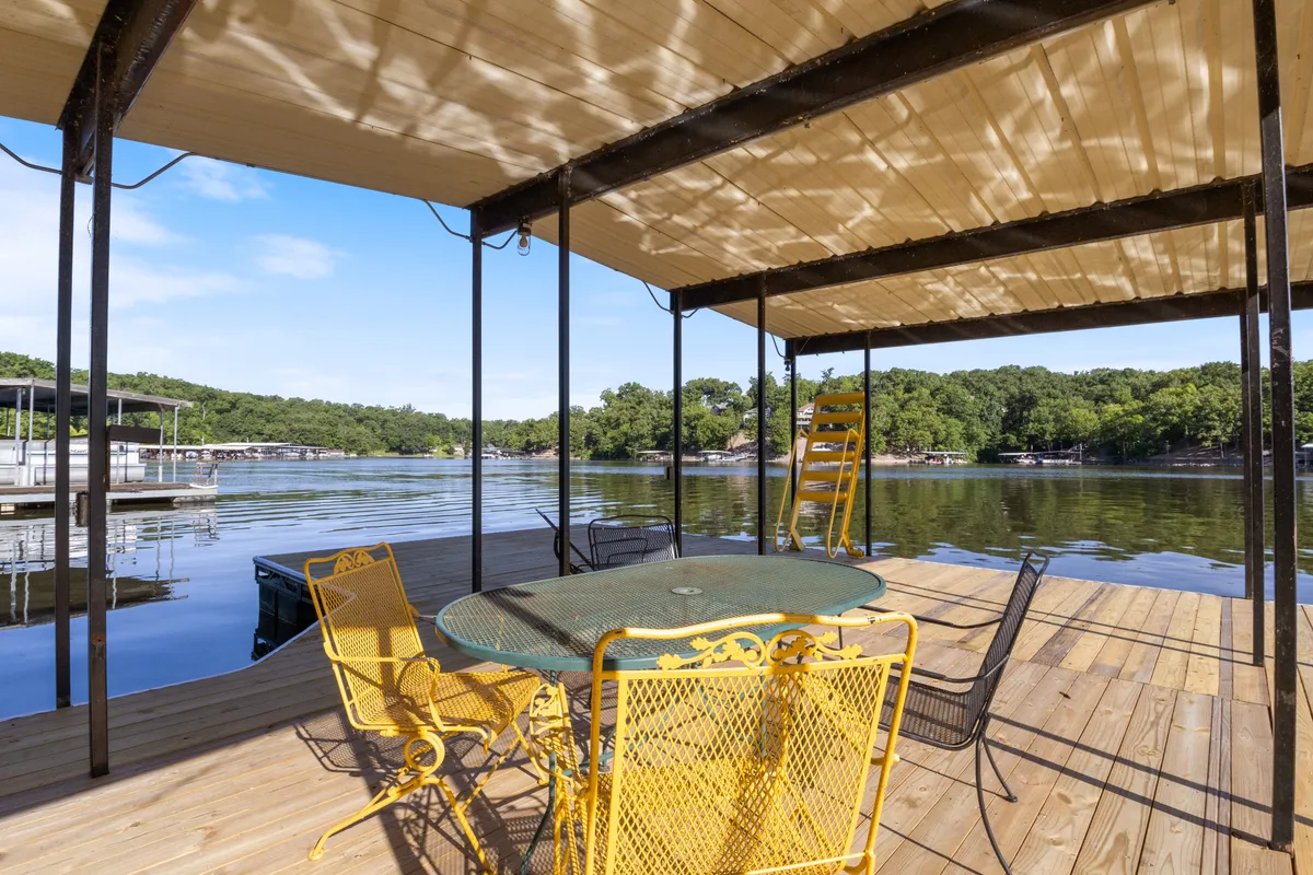 A lakeside setup featuring yellow chairs and a metal table overlooking a serene lake, with boathouses visible in the background. The scene is bright and tranquil, ideal for relaxed outdoor dining or enjoying drinks with friends at the water’s edge.