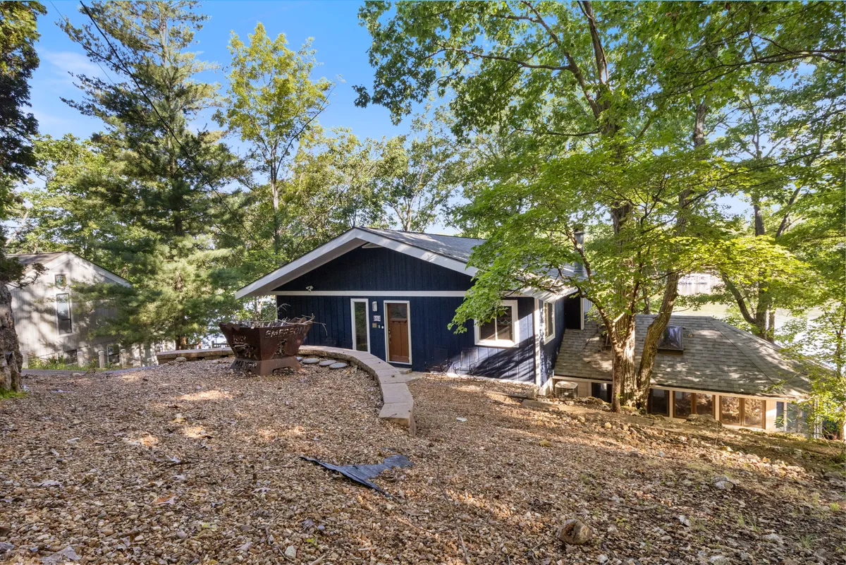 A cozy outdoor fire pit area sits in front of a home with dark siding, with a pathway leading from the house to the gathering space. Lush greenery surrounds the scene, evoking a relaxed and inviting atmosphere for evenings outdoors.