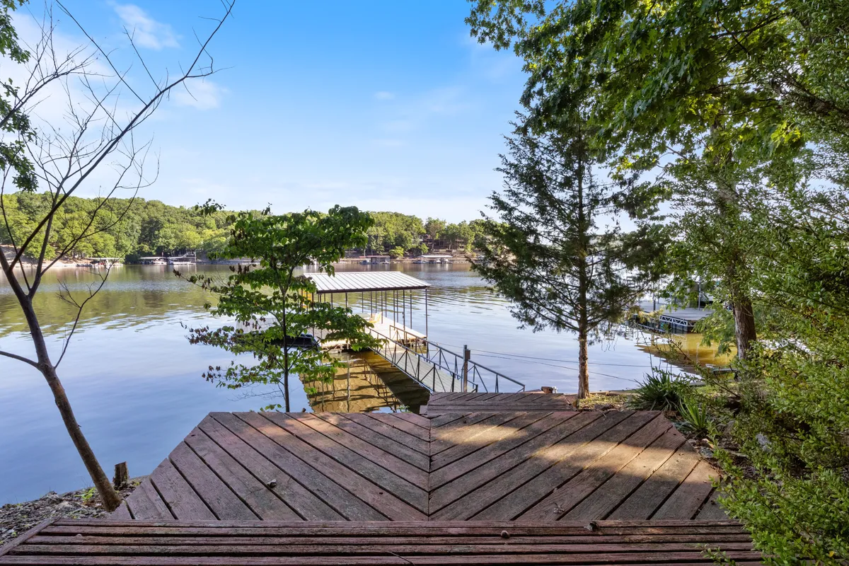 A tranquil wooden dock extends over calm lake waters, surrounded by lush trees. The serene setting features clear reflections on the water and the dock provides easy access for boating, fishing, or simply unwinding next to the lake.