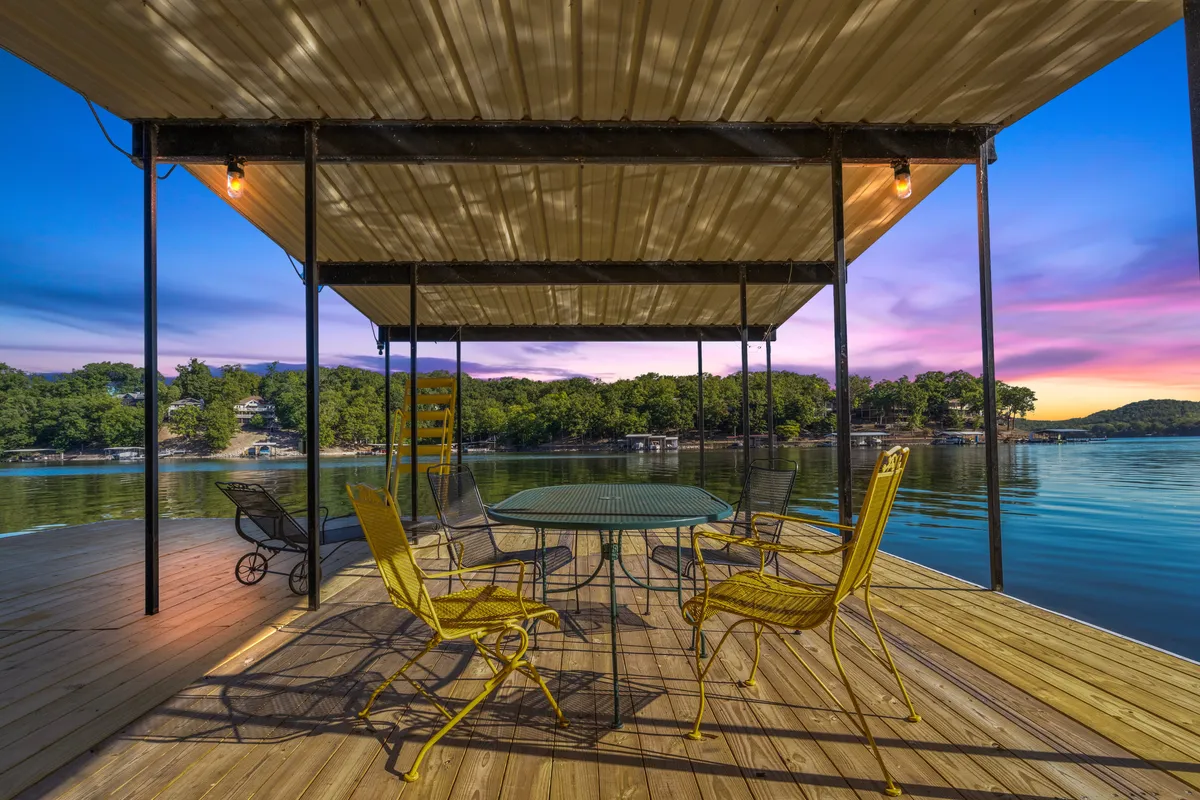 The image showcases the exterior of a lake house patio with bright yellow chairs around a round table, overlooking calm lake waters. The sky is painted in warm sunset hues, reflecting off the water. The setting suggests a peaceful, welcoming spot for dining, relaxing, or gathering with friends and family.