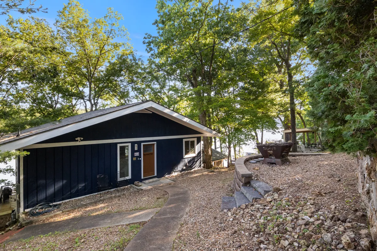 A peaceful outdoor scene highlights a firepit area with seating, surrounded by trees and overlooking the lake. The blue house peeks through the greenery, and the space is set up for gatherings, s’mores, and evening relaxation.